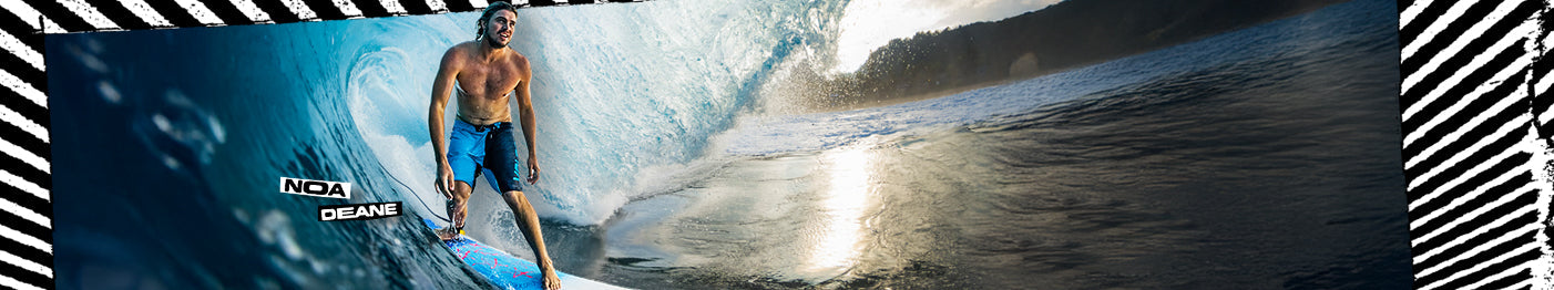 A surfer rides inside a large wave near the shore at sunset. Text on the image reads Noa Deane. The scene is bordered by a black-and-white striped pattern.