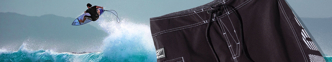 A surfer catches air above a wave, while a close-up of black board shorts with white stitching appears in the foreground.