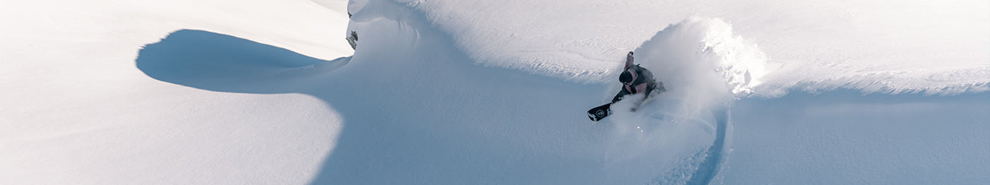 A snowboarder carves through deep, fresh powder on a snowy mountain slope, creating a spray of snow in the bright sunlight.