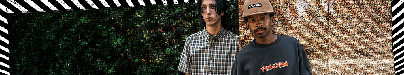 2 men standing in front of a wall, with a plaid short sleeve and graphic tee