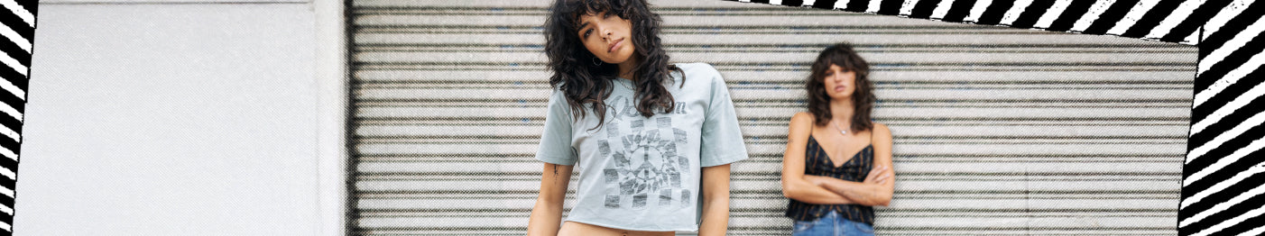 Two women stand in front of a metal shutter; one in front wearing a light crop top and beige pants, the other in the background wearing a black top and jeans, both with wavy hair. Striped border frames the image.