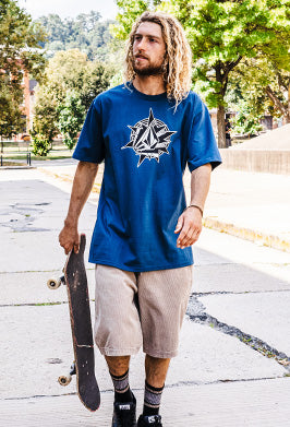Man holding a skateboard wearing a blue t-shirt with a graphic design outdoors.