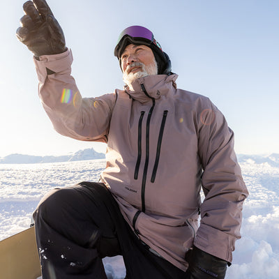 Person in a light brown snow jacket with black accents, sitting on a snowy landscape.