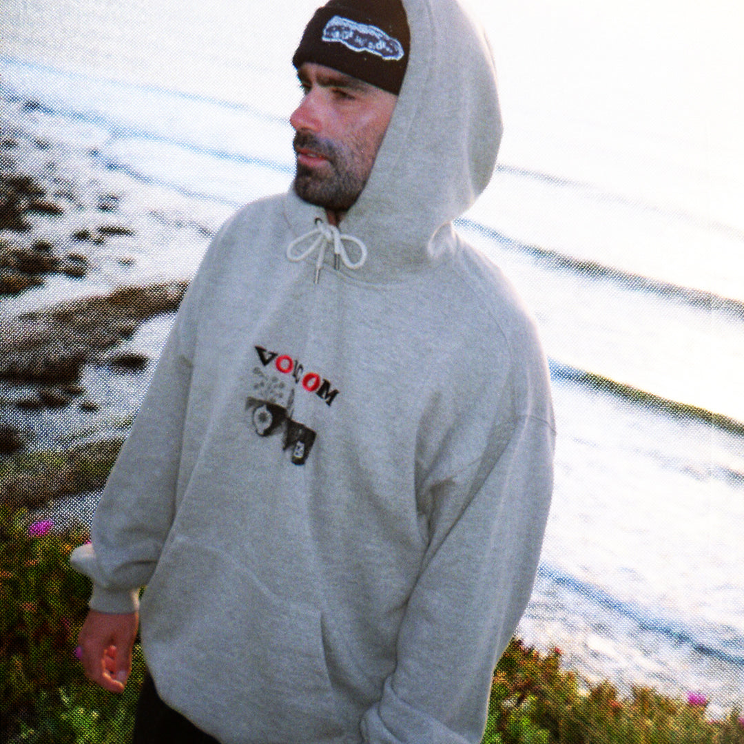 Man wearing a gray hoodie with a logo, standing outdoors near water.