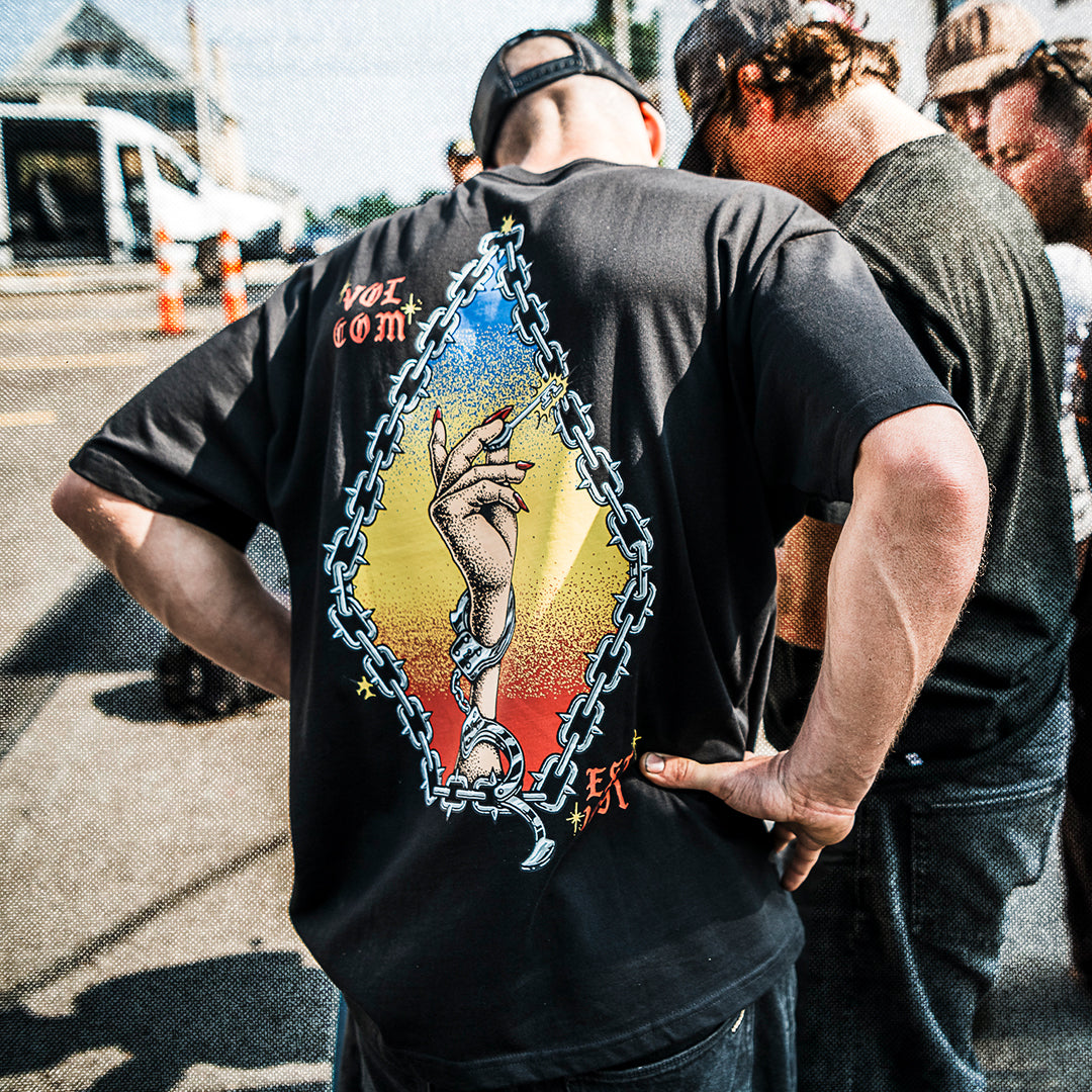 Person wearing a black t-shirt with a graphic design, standing outdoors with other people in the background.