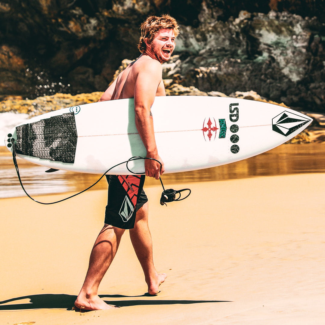 Man walking on a beach holding a surfboard with a rocky background