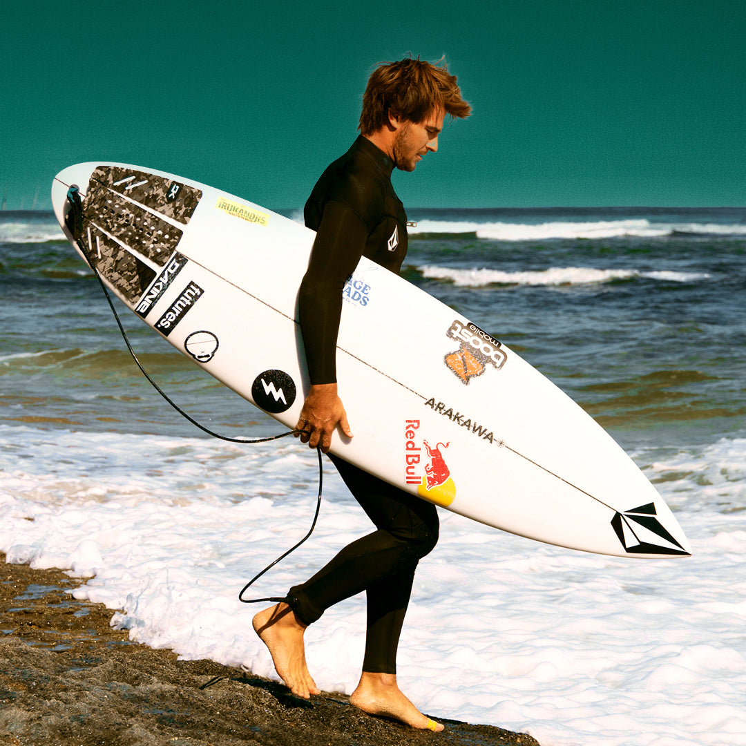 Person in a wetsuit holding a surfboard on a beach with ocean waves in the background