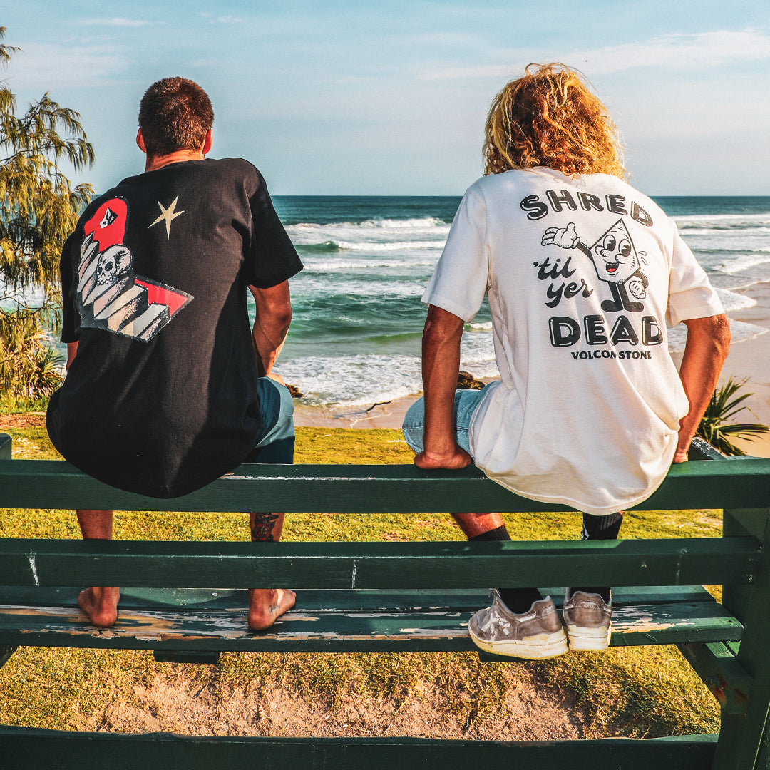 Two people sitting on a bench by the ocean, wearing t-shirts with visible text and graphics.