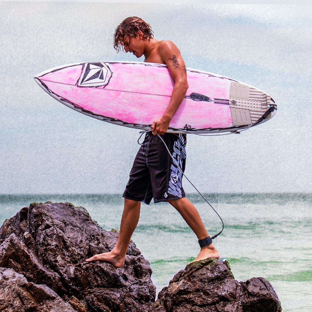 Person holding a pink surfboard on rocks with ocean in the background