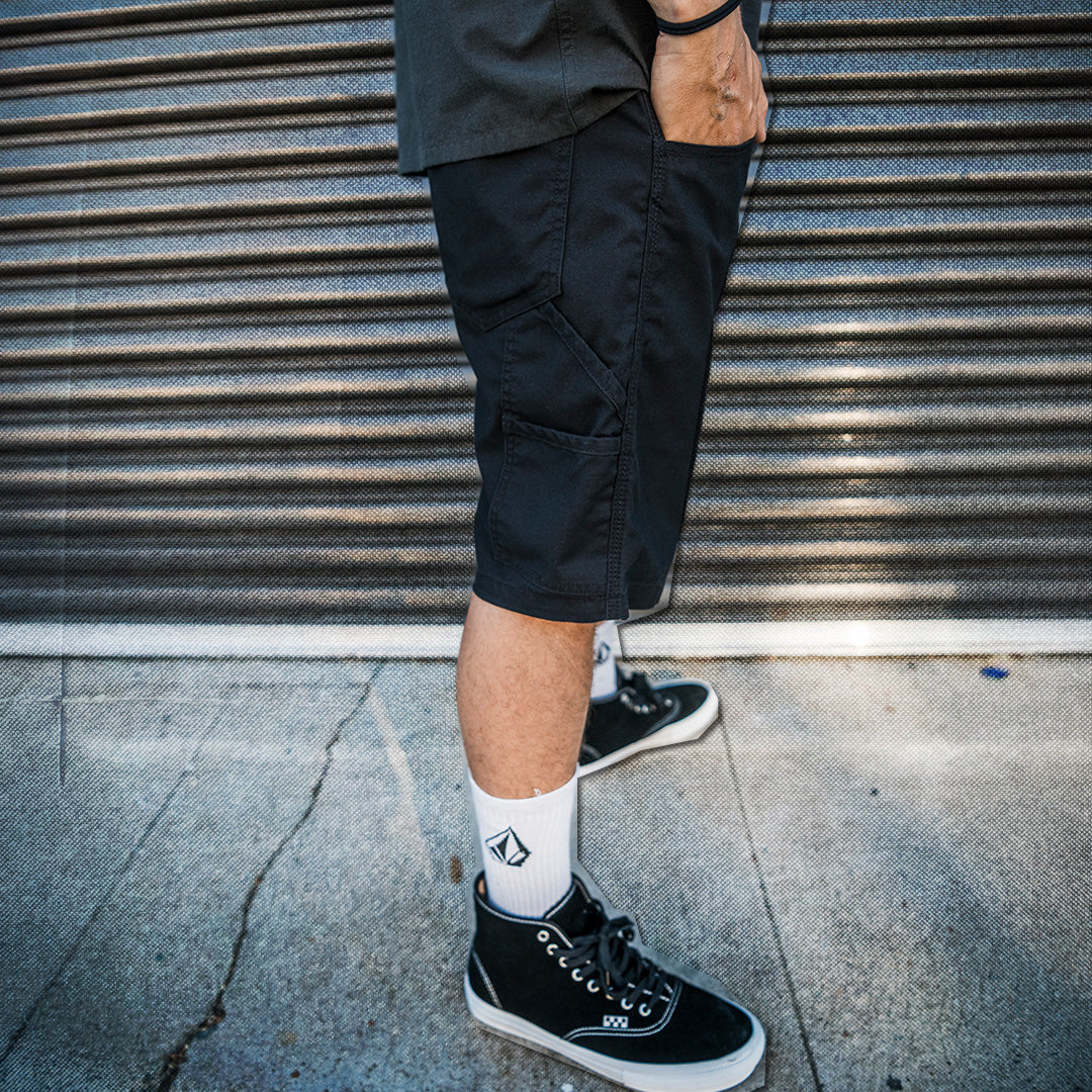 Person wearing black shorts and black sneakers with a white sock, standing against a metal shutter.