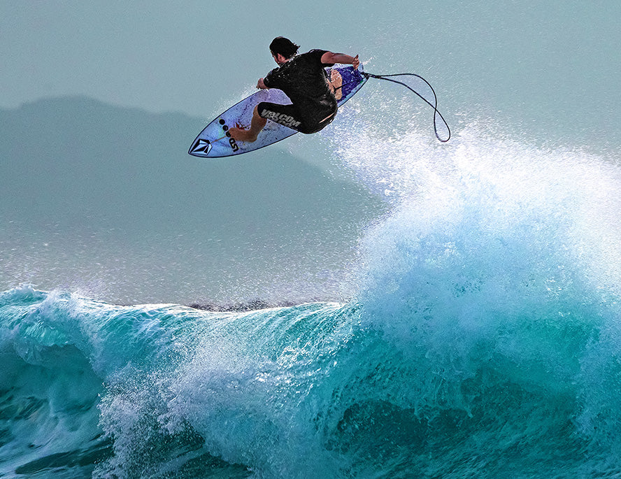 A surfer catches air above a turquoise wave, holding onto a white surfboard with a leash trailing behind. The ocean spray splashes dramatically beneath him against a hazy blue sky.