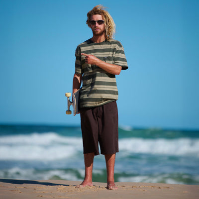 Man with a striped shirt and sunglasses holding a skateboard on a beach.