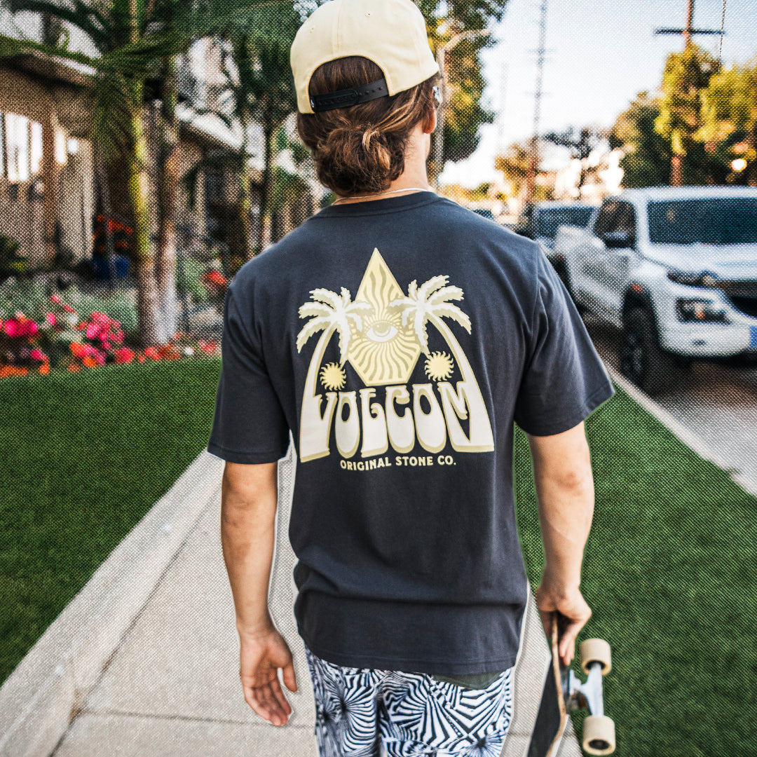Person in white cap and dark shirt with Volcom branding carrying skateboard and walking on street