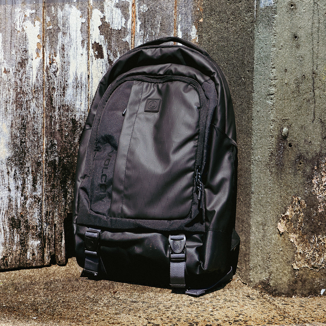 A black technical backpack with multiple zippered compartments and side compression straps, leaning against a weathered wooden and concrete wall.