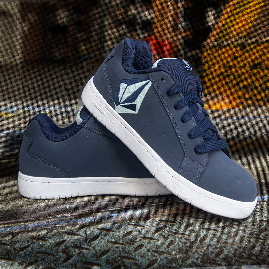 A pair of Volcom low-top sneakers in navy blue suede and canvas with white soles, displayed on a textured concrete surface with an industrial background.
