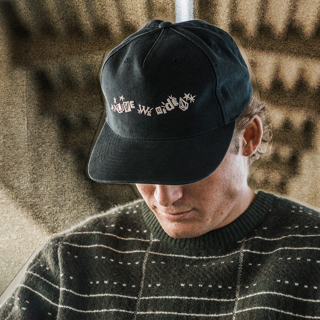 A man wearing a black baseball cap with white embroidered text that reads 'ALIVE WE RIDE?' and a dark striped sweater, shown in a close-up shot.