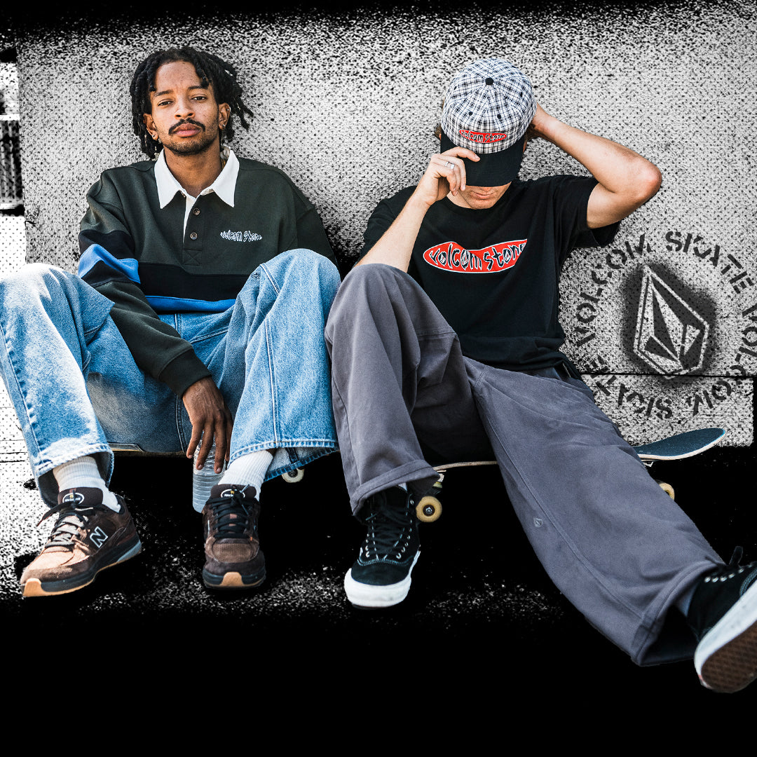 Two men sitting on skateboards against a graffiti-covered wall, wearing New Balance sneakers, with one wearing a black and blue striped polo and the other a black graphic t-shirt and gray sweatpants.