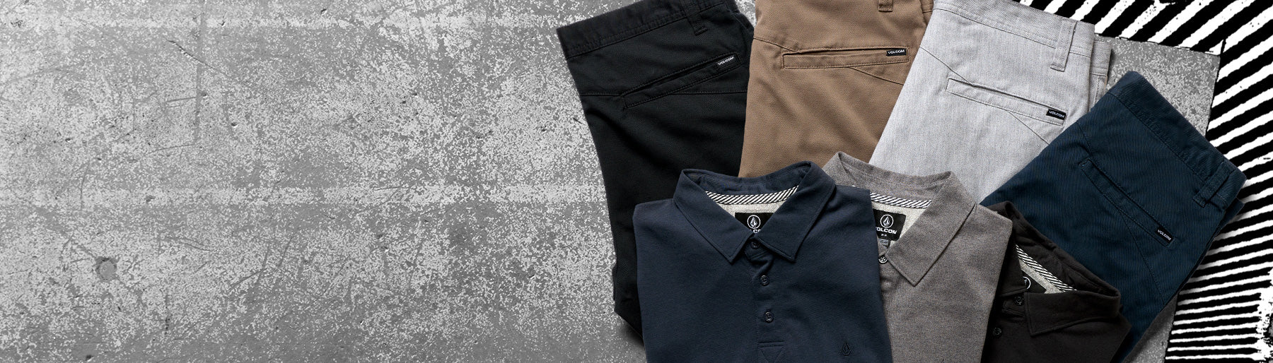 A styled flat-lay of folded men's clothing on a textured gray concrete background, including dark navy, black, tan, light gray, and dark teal shirts and trousers, alongside a black-and-white striped fabric accent.