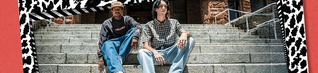 Two young men sit on stone steps outside a building, one wearing a cap and dark sweatshirt, the other in a plaid shirt. The image is bordered by a black-and-white patterned graphic frame.