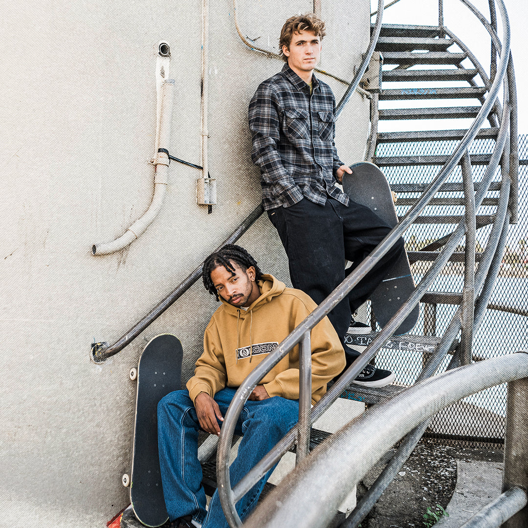 Two young men posing on an outdoor spiral metal staircase against a concrete wall, each holding a skateboard. One is wearing a black and gray plaid flannel shirt, black pants, and white sneakers, while the other is wearing a tan hoodie with a graphic print and blue jeans.