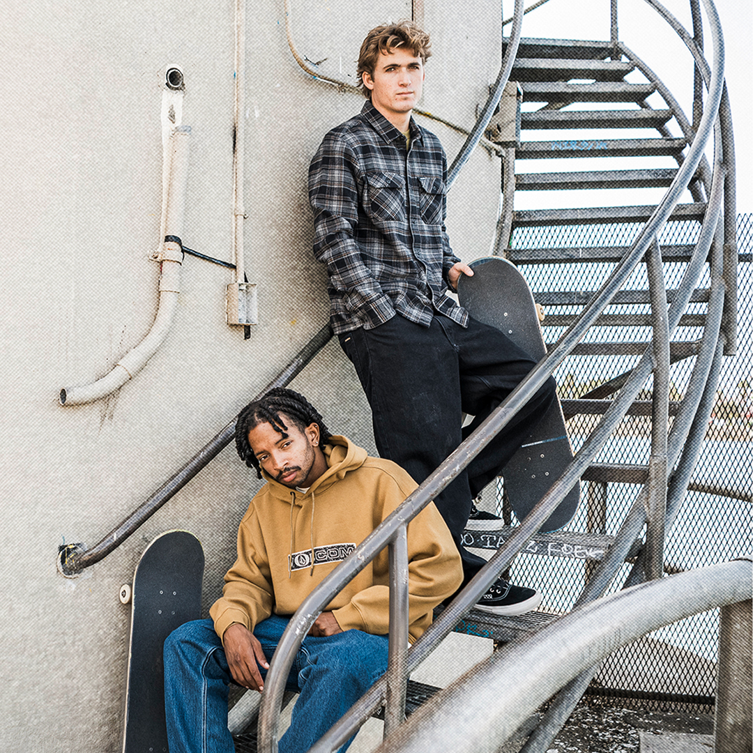 Two skateboarders posing on a metal spiral staircase against a concrete wall. The skateboarder on the top step wears a dark gray plaid flannel shirt and black pants, holding a skateboard.