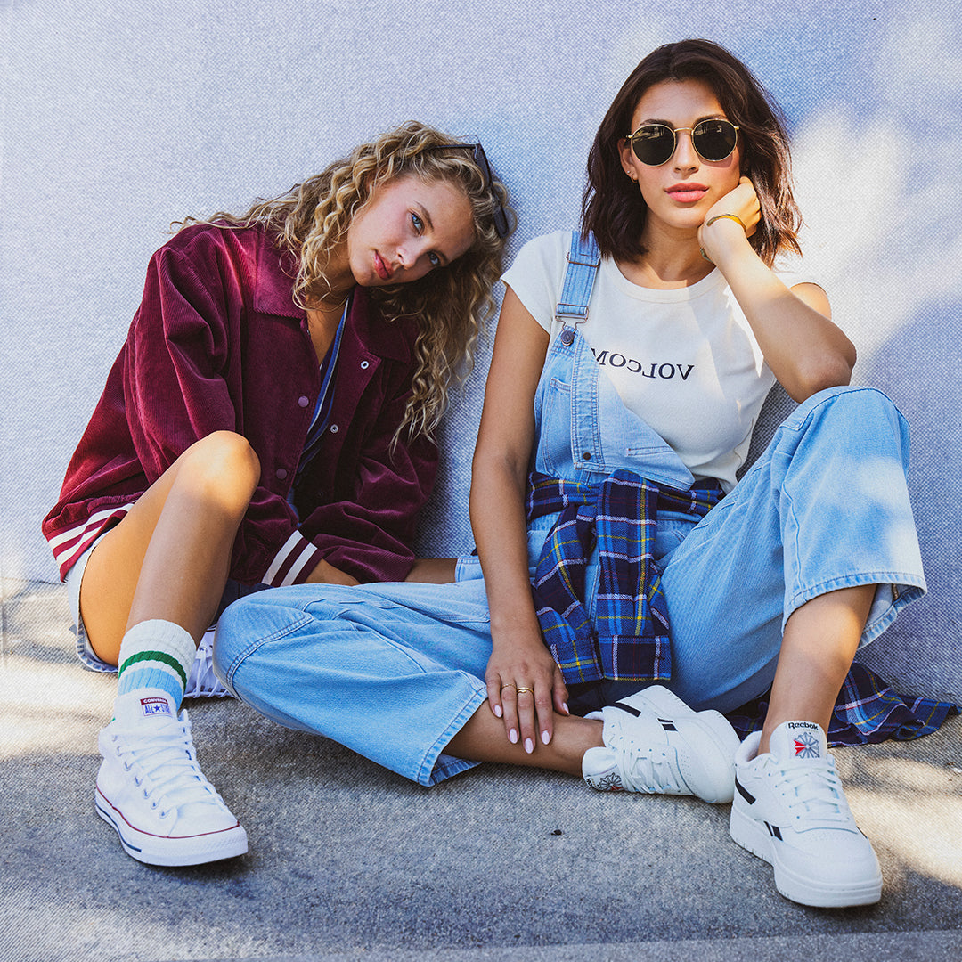 Two women sitting on concrete outdoors wearing casual streetwear. One wears a burgundy velvet overshirt, denim shorts, and white sneakers.