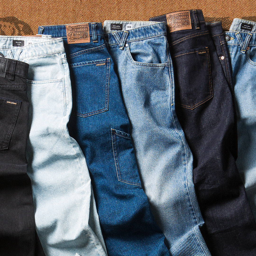 Six pairs of jeans in different denim washes arranged side by side on a brown textured surface. The washes include black, very light blue, medium blue, light blue, dark navy, and medium blue.