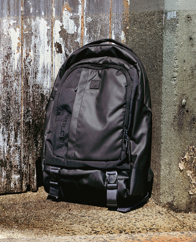 A black Volcom backpack made of durable nylon, leaning against a weathered wooden and concrete wall.