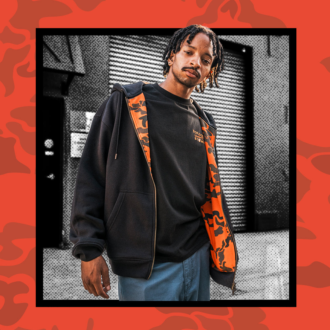 A man wearing a black zip-up hoodie with bright orange patterned lining, layered over a black t-shirt and blue jeans, set against a stylized black-and-white urban background with a red patterned border.