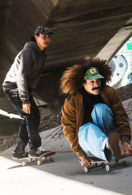 Two skateboarders under a concrete bridge. One person stands on a skateboard wearing a dark gray hoodie, black pants, and a black cap.