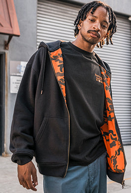 A man wearing a black zip-up hoodie with a bright orange camouflage-patterned lining over a black t-shirt and blue jeans, standing in front of a gray metal shutter.