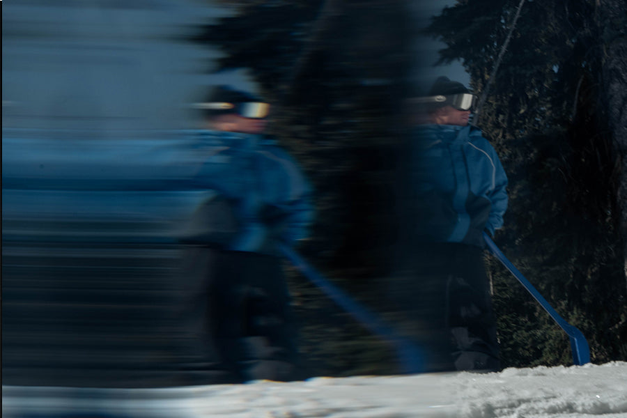 Two people in blue winter gear and goggles skiing or snowboarding through deep snow, with dark trees and a blurred outdoor winter background.