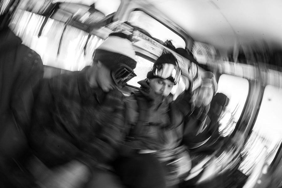 Black and white action photograph of a person wearing a knit beanie and goggles, seated inside a moving vehicle with motion blur.