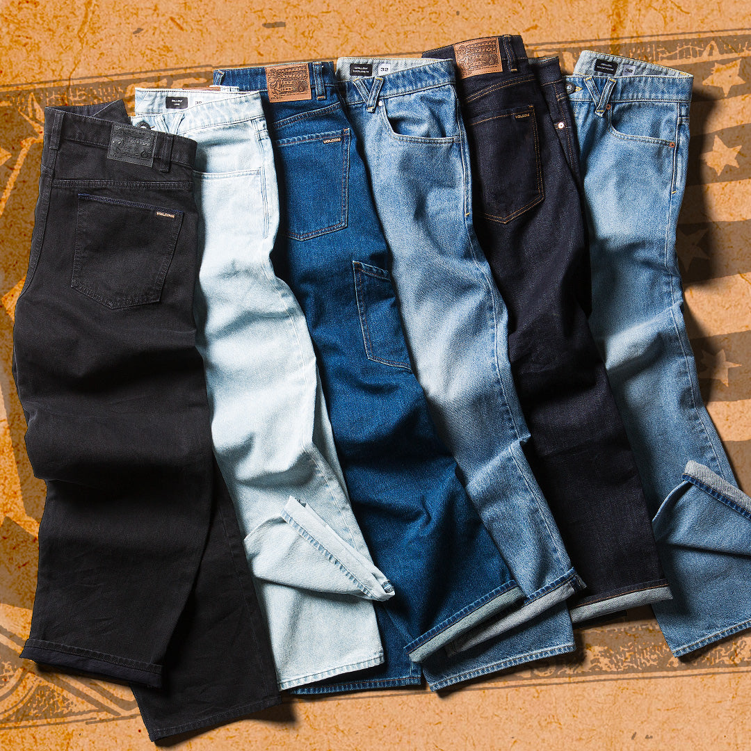 Six pairs of folded denim jeans in different washes are arranged side by side on a textured tan surface. The jeans include black, very light blue, medium blue, dark blue, and light blue washes.
