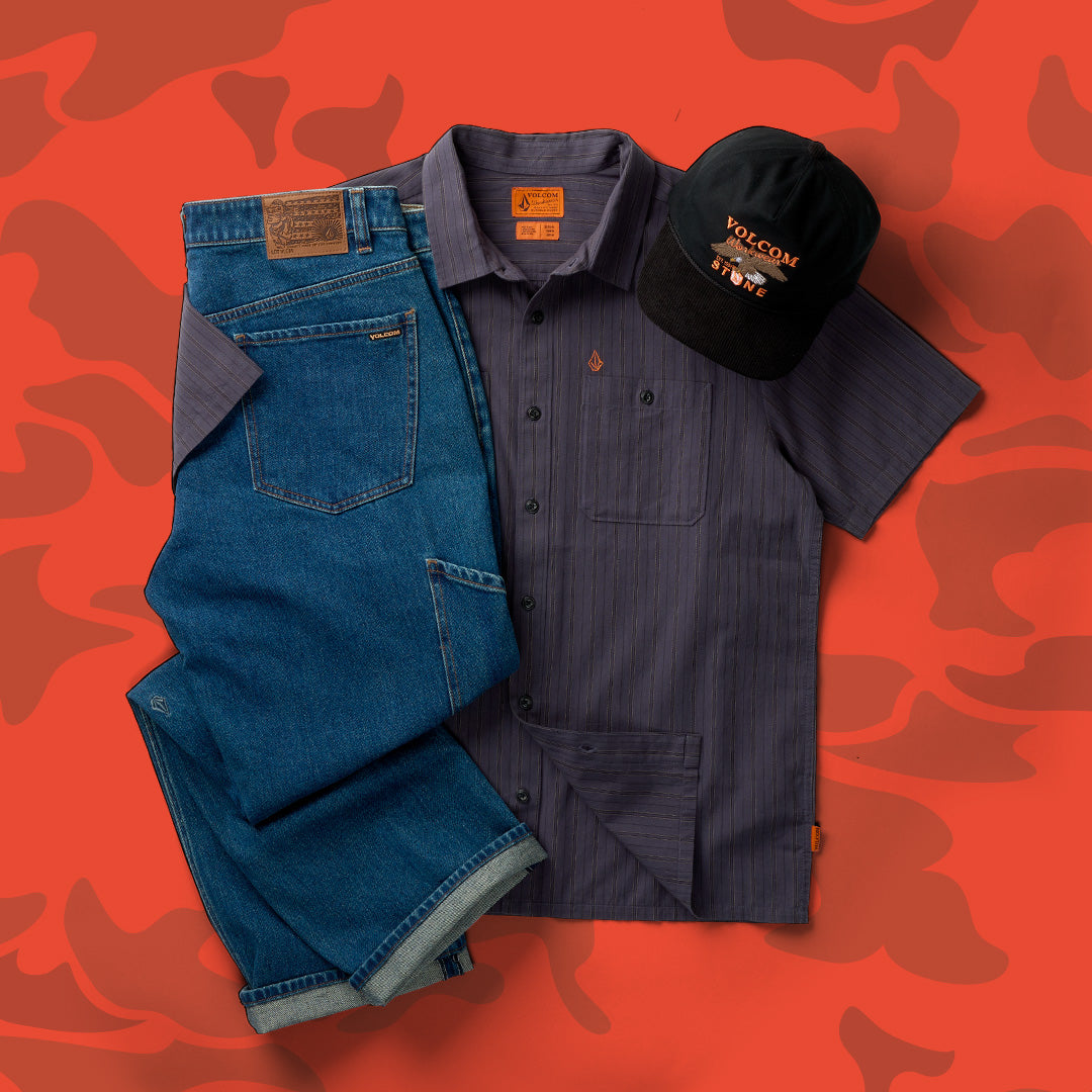 A styled flat-lay of a Volcom apparel set featuring a dark gray pinstriped short-sleeve button-up shirt, blue denim jeans, and a black Volcom cap, arranged on a red camouflage patterned background.