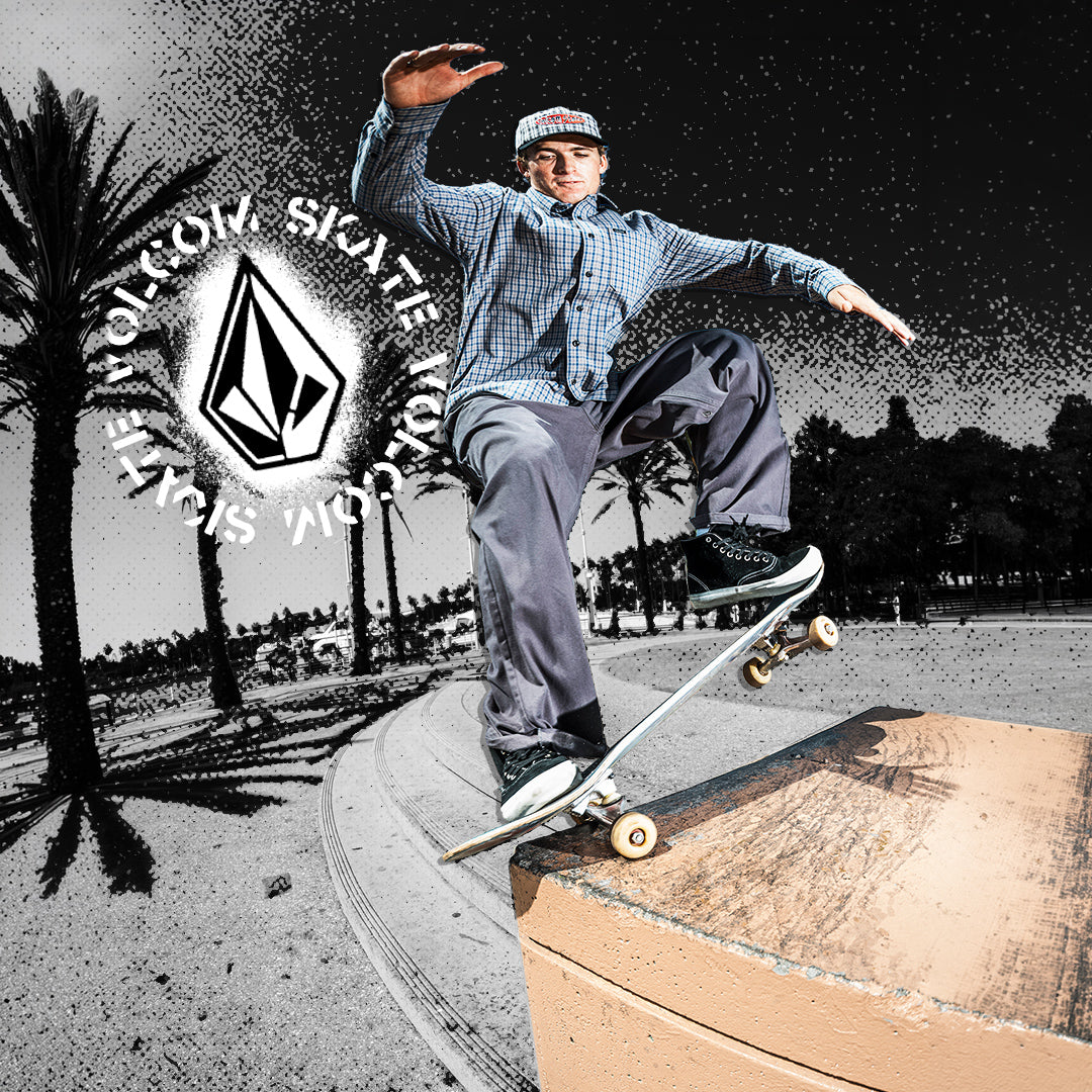 A skateboarder in a blue plaid shirt and gray pants performs a trick on a ramp, with a large Volcom Skateboard logo and palm trees in the background.