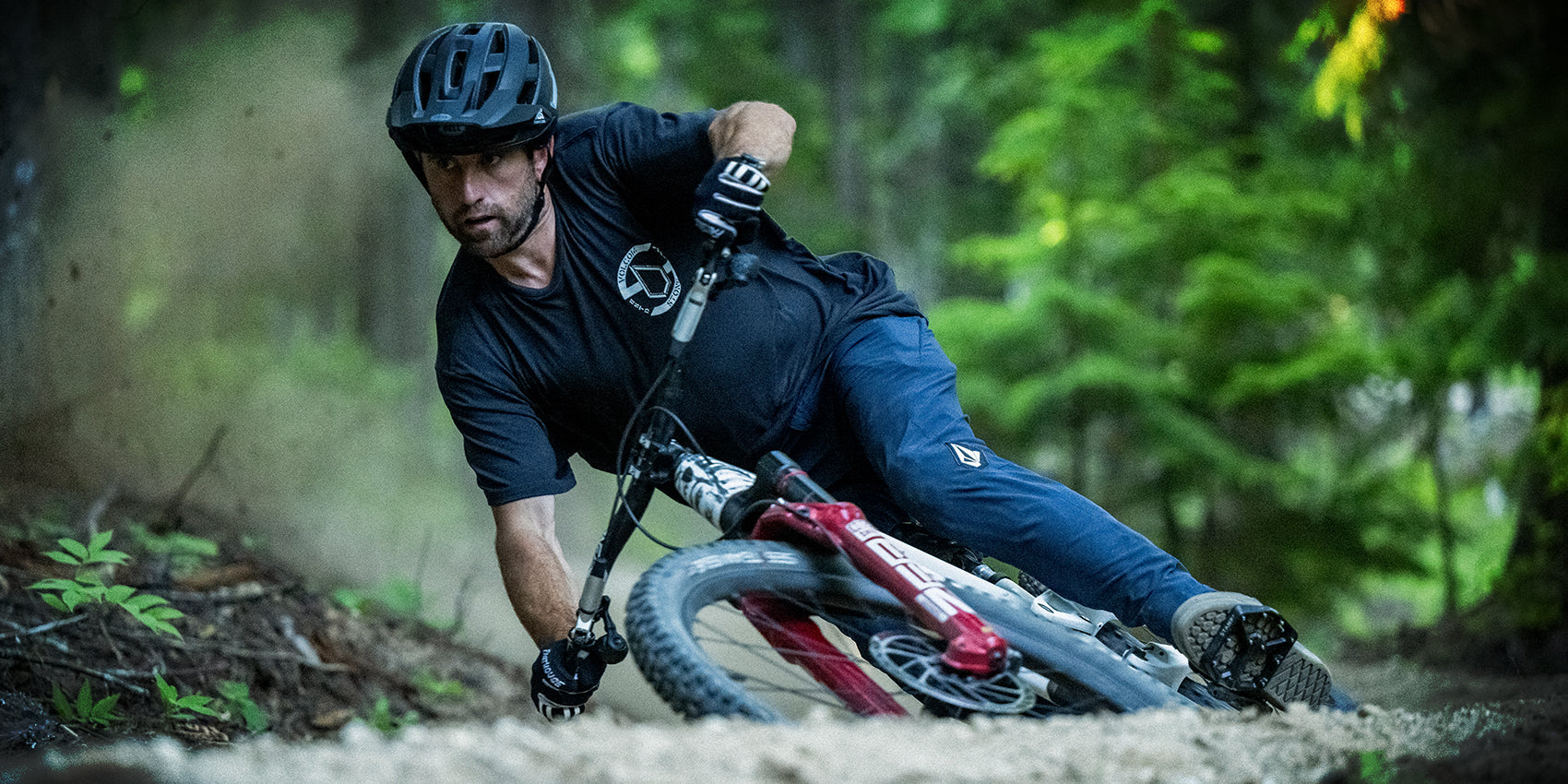 A mountain biker wearing a helmet and dark clothing leans sharply into a turn on a dirt trail through a forest, kicking up dust and surrounded by green foliage.