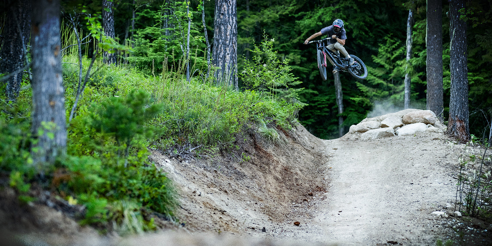 A mountain biker wearing a helmet and goggles jumps off a dirt ramp on a forest trail, surrounded by tall trees and green foliage. Dust trails behind the bike, highlighting the dynamic motion.