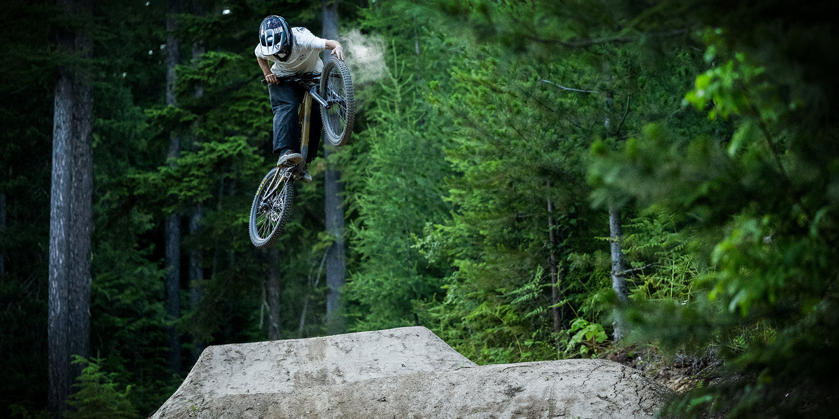 A mountain biker wearing a helmet performs a jump and trick in the air on a dirt trail surrounded by dense green forest.
