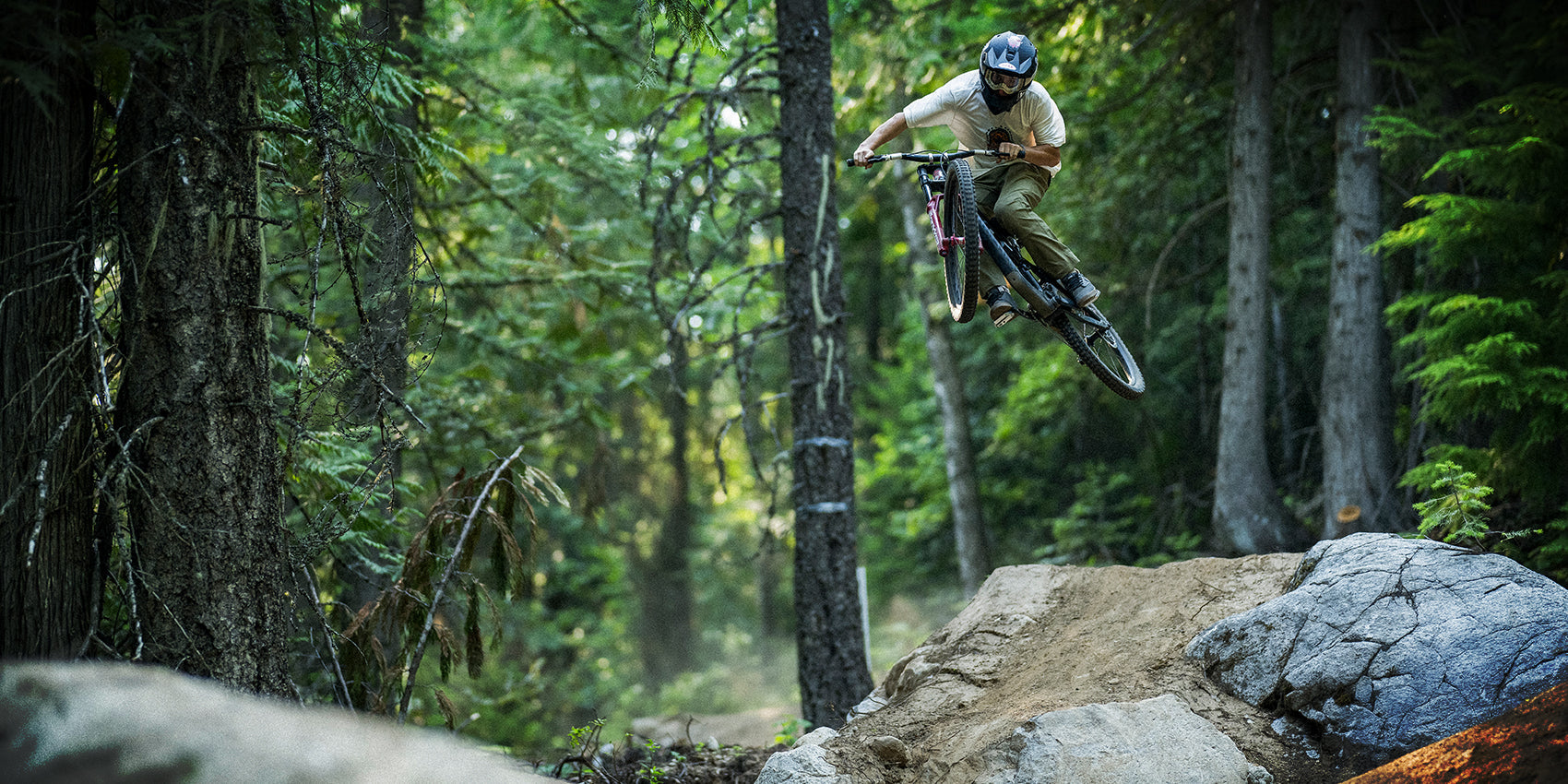 A mountain biker wearing a helmet and goggles jumps off a dirt ramp over rocks in a dense, green forest. Sunlight filters through the trees as the rider is mid-air, displaying skill and control.