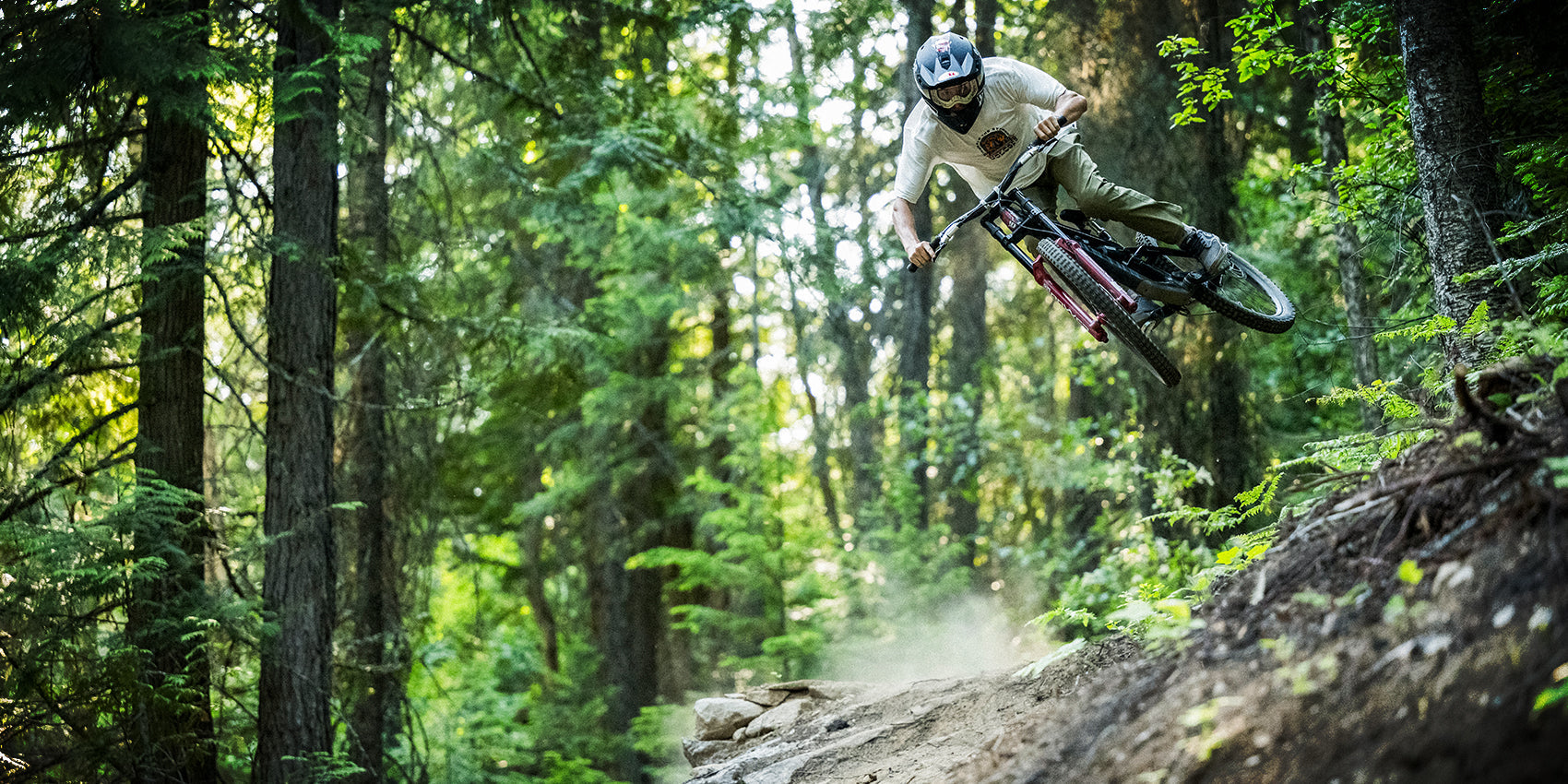 A mountain biker wearing a helmet and goggles rides at high speed through a forest trail, kicking up dust while leaning sharply to the side. Tall green trees and dense foliage surround the dirt path.