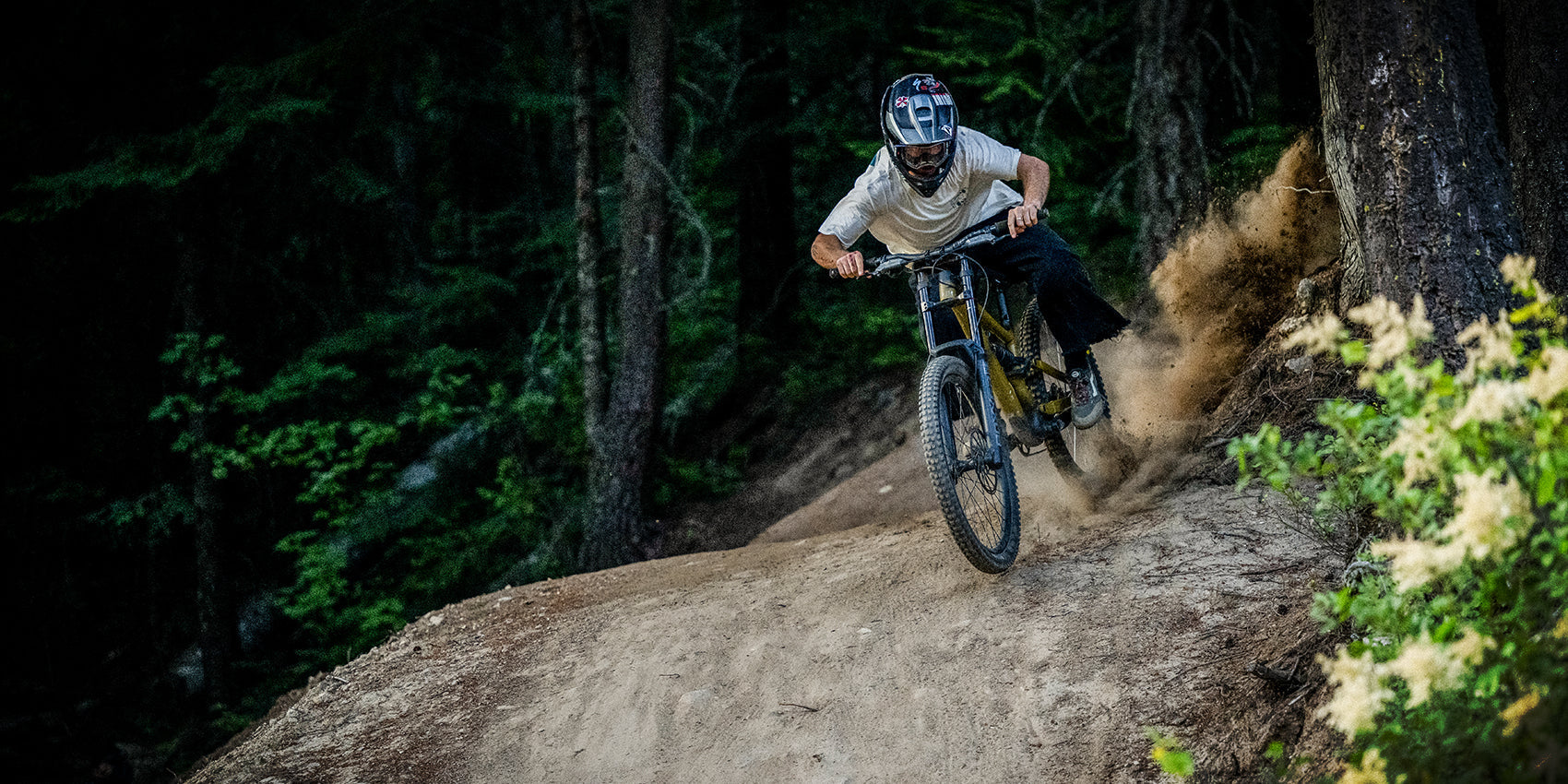 A mountain biker wearing a helmet rides downhill on a dirt trail in a forest, kicking up dust as they speed around a turn.