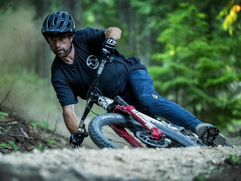A mountain biker in a black helmet and dark clothing leans sharply into a turn on a dirt trail in a forest, kicking up dust as he rides.