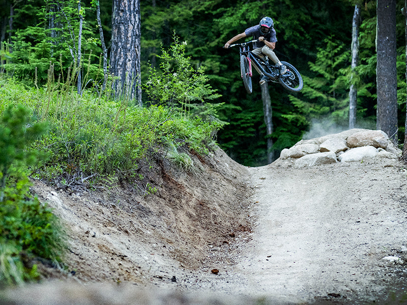 A mountain biker wearing a helmet jumps off a dirt ramp on a forest trail, with trees and greenery in the background and a cloud of dust behind the bike.