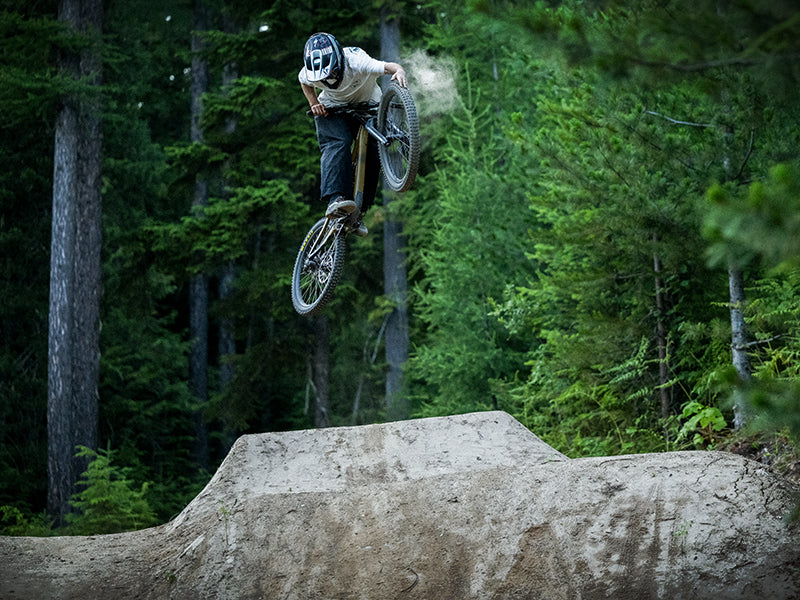 A mountain biker wearing a helmet performs a jump trick in mid-air over a dirt ramp, surrounded by dense green forest.