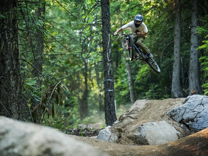 A mountain biker wearing a helmet jumps off a dirt ramp in a forested trail, surrounded by trees and rocks, with sunlight filtering through the leaves.