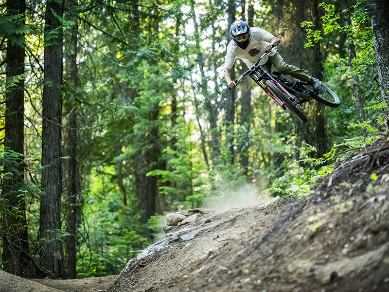 A mountain biker wearing a helmet and light-colored clothing jumps off a dirt trail in a forest, leaning sideways in mid-air with trees and greenery in the background.