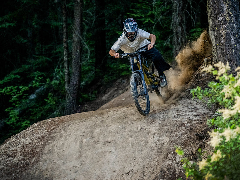 A mountain biker wearing a helmet rides downhill on a dirt trail through a forest, kicking up dust as they take a sharp turn near trees.