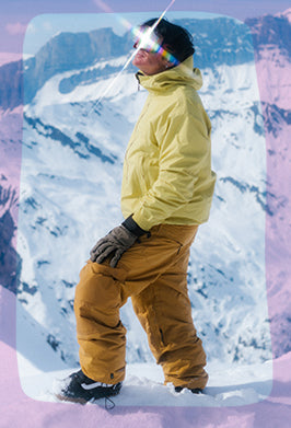 A person standing in snow wearing a bright yellow ski jacket, tan snow pants, black gloves, and black ski boots, with a snowy mountain backdrop and a pink-toned border effect.