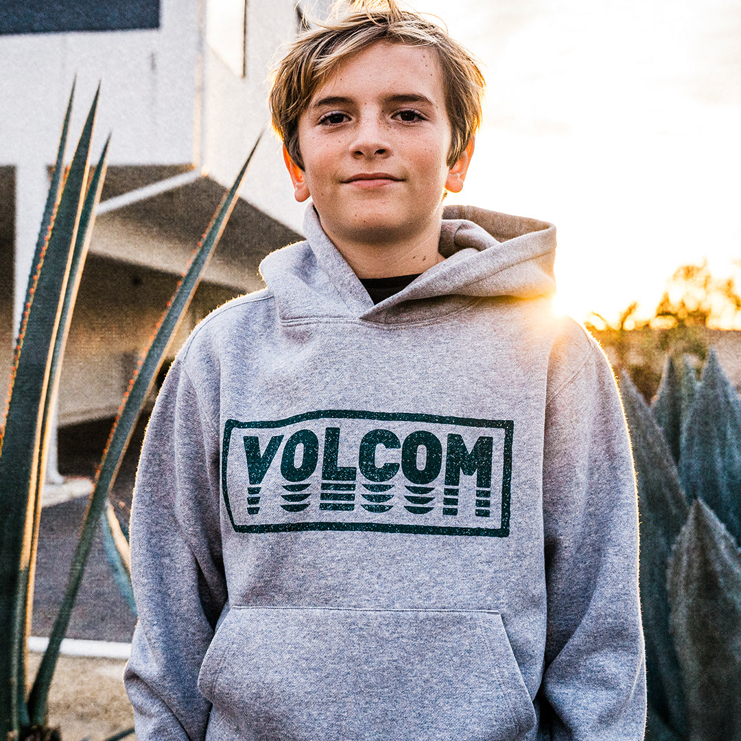 A young man wearing a heather gray Volcom hoodie with a dark green graphic logo on the chest, standing outdoors in front of a building with plants and warm sunset light.
